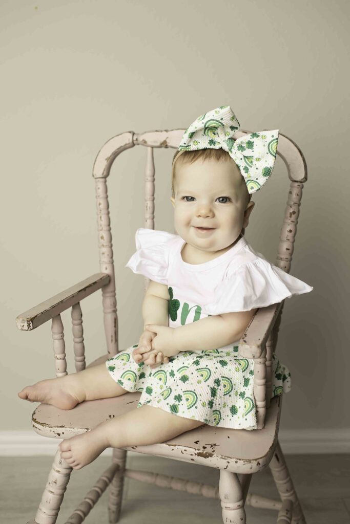 Captured in a whimsical Lucky Clover theme, the one-year-old birthday girl poses sweetly in a wicker chair at Chunky Monkey Photography Studio in Azle, TX