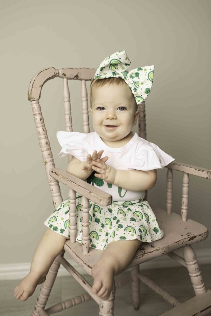 The birthday girl in a green dress sits happily in a wicker chair surrounded by Lucky Clover decorations at Chunky Monkey Photography Studio in Azle, TX