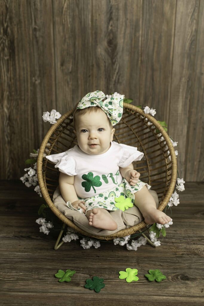 The birthday girl, dressed in green, smiles as she sits in a wicker chair amidst the Lucky Clover decorations at Chunky Monkey Photography Studio in Azle, TX