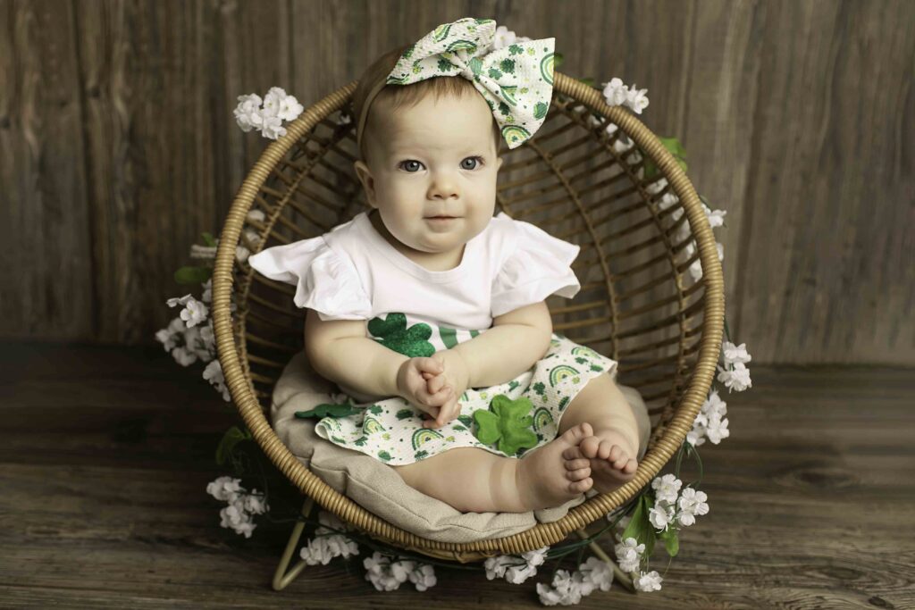 The birthday girl in a green dress sits happily in a wicker chair surrounded by Lucky Clover decorations at Chunky Monkey Photography Studio in Azle, TX