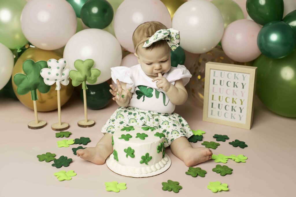 A one-year-old in a green dress enjoys her cake smash surrounded by Lucky Clover decor at Chunky Monkey Photography Studio in Azle, TX, celebrating her first birthday