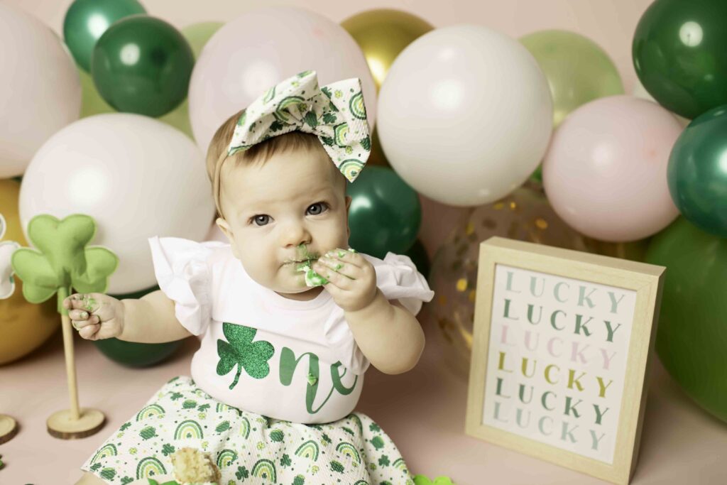 A joyful one-year-old in a green dress digs into her cake during a Lucky Clover-themed cake smash at Chunky Monkey Photography Studio in Azle, TX