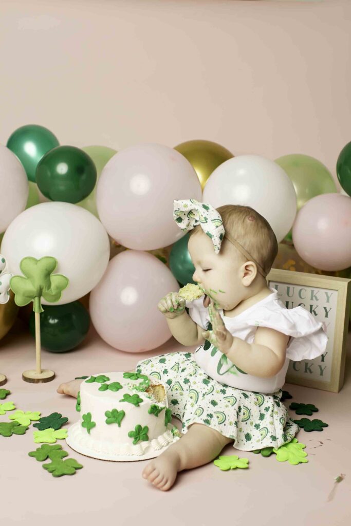 The birthday girl in a green dress laughs as she smashes her cake amidst the Lucky Clover theme at Chunky Monkey Photography Studio in Azle, TX