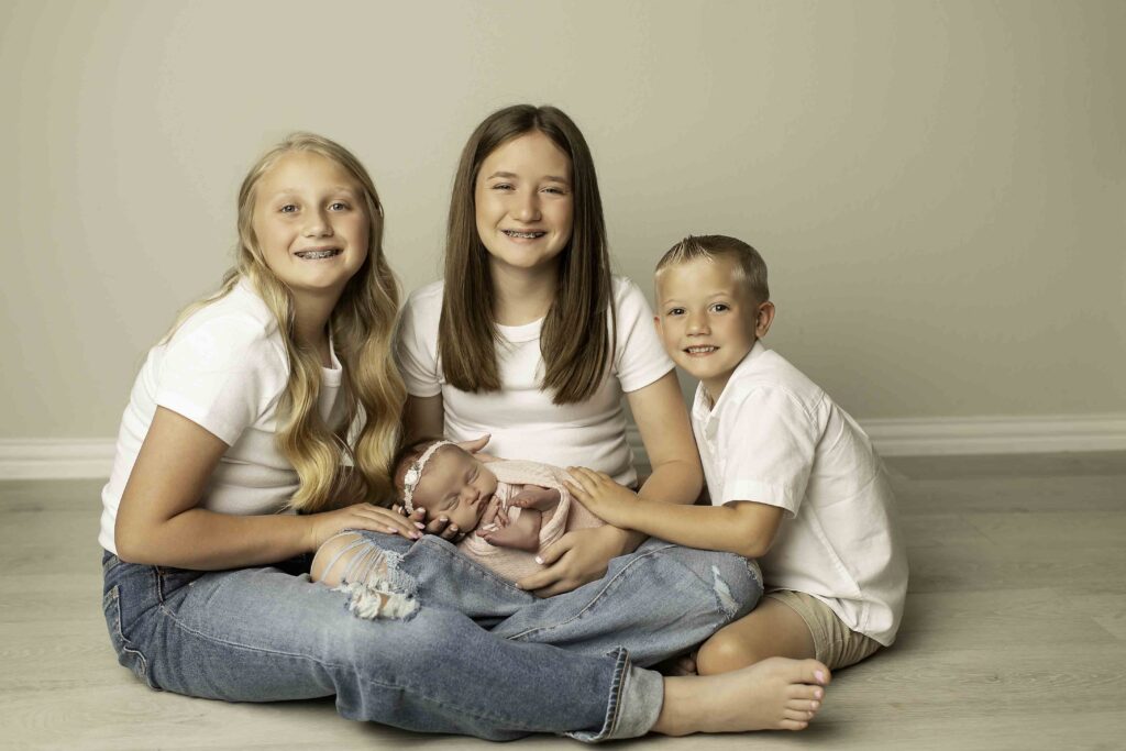 A heartwarming moment with three siblings lovingly surrounding their newborn sister, sharing smiles and excitement, beautifully captured by Chunky Monkey Photography.