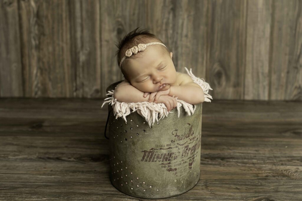 A newborn girl peacefully resting in a rustic fishing bucket, surrounded by soft blankets, captured in a sweet and serene moment by Chunky Monkey Photography