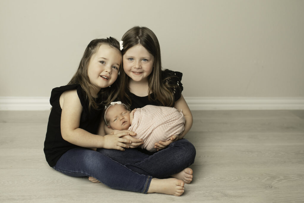 A heartwarming family moment with two big sisters and their newborn baby sister, sharing love and joy in this precious photo by Chunky Monkey Photography