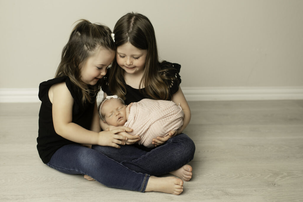 Two proud big sisters smile in delight as they hold their newborn baby sister close, capturing the love and bond between them, beautifully shot by Chunky Monkey Photography