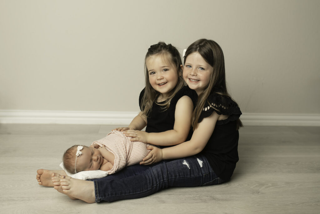 A beautiful moment captured with two big sisters lovingly surrounding their newborn baby sister, full of joy and excitement, shot by Chunky Monkey Photography