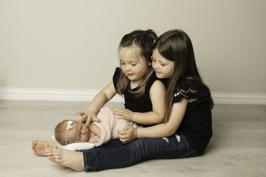 Big sisters gaze lovingly at their newborn sister, capturing the warmth and excitement of welcoming a new family member, shot by Chunky Monkey Photography.