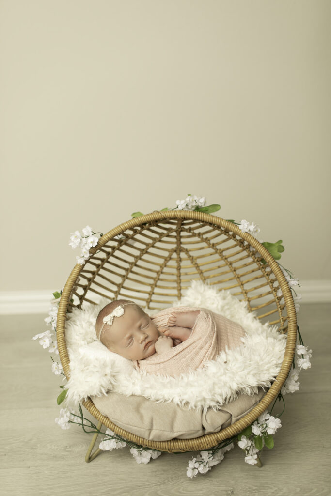 A newborn girl curled up comfortably in a wicker chair, captured in a serene and timeless pose by Chunky Monkey Photography.