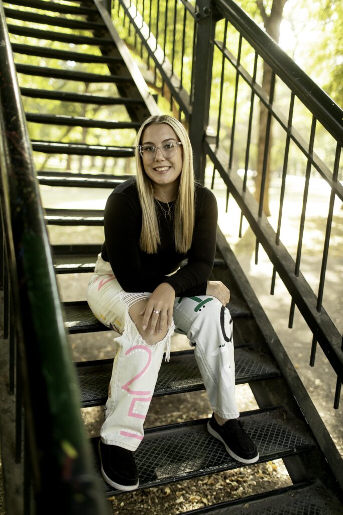 A senior girl smiles warmly as she poses with the urban landscape of Fort Worth's Trinity Park in the background during her photoshoot