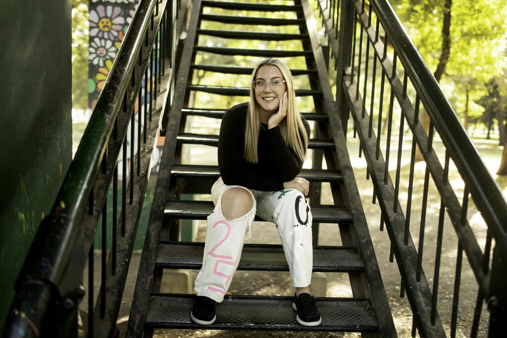 A senior girl strikes a confident pose on metal stairs in an urban area, with the cityscape of Fort Worth in the background during her photoshoot