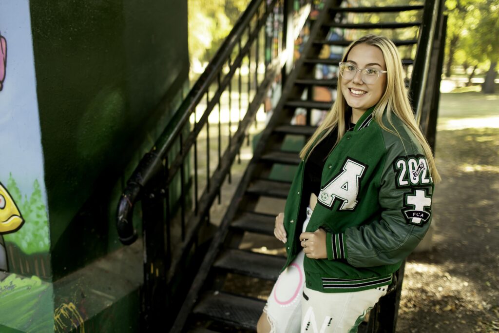 A senior girl leans on metal stairs in the heart of an urban area, showcasing her unique style with the Fort Worth cityscape behind her