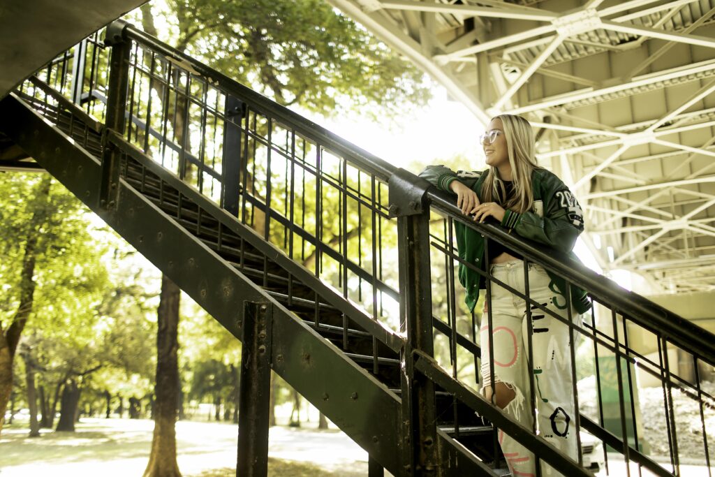 Captured on industrial metal stairs, a senior girl stands proudly in her graduation attire, with the urban Fort Worth skyline adding a modern touch