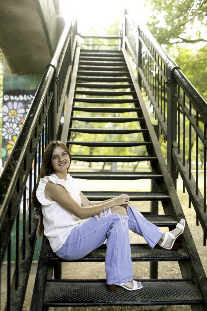 Senior girl sitting casually on metal stairs, with a natural smile in her white shirt and blue jeans