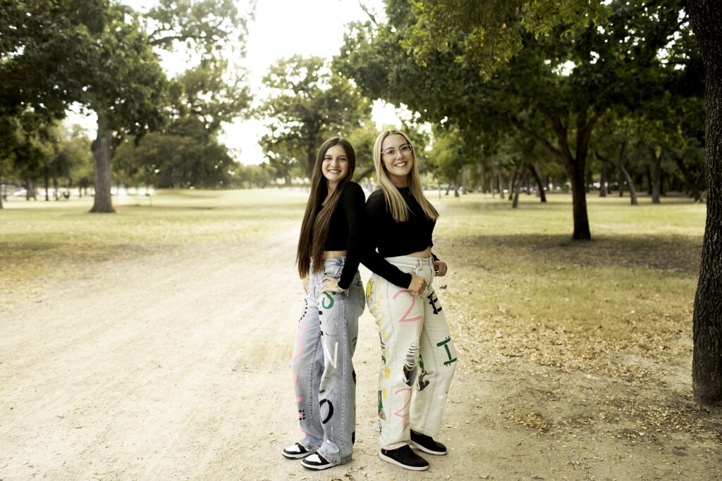 Two senior girls pose together in their 2025 senior jeans, laughing and enjoying the moment in an urban area of Fort Worth, with the city skyline behind them