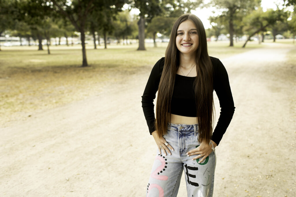 Senior girl smiling brightly on a dirt road at Trinity Park, wearing a black shirt and 'Senior 2025' pants