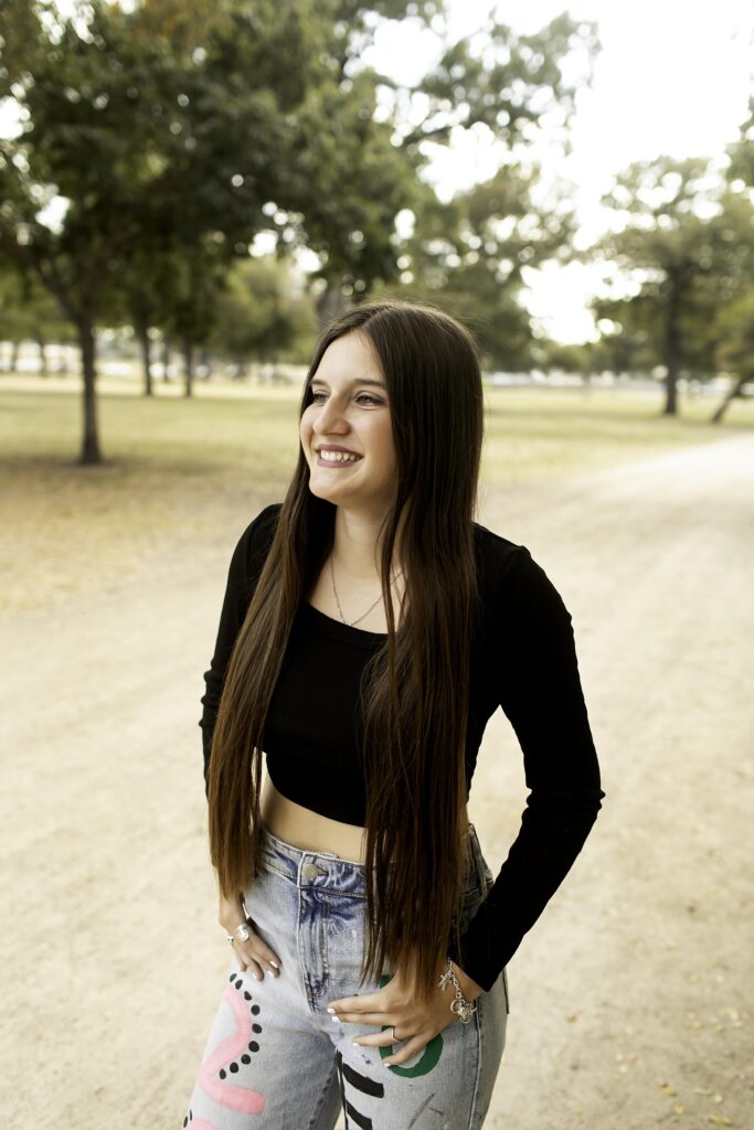 Senior girl posing with a relaxed smile on a dirt road at Trinity Park, showcasing her black shirt and 'Senior 2025' pants