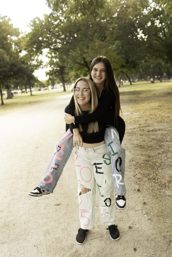 Two senior girls stand side by side in their 2025 senior jeans, smiling confidently in an urban Fort Worth setting, with the city’s skyline in the background