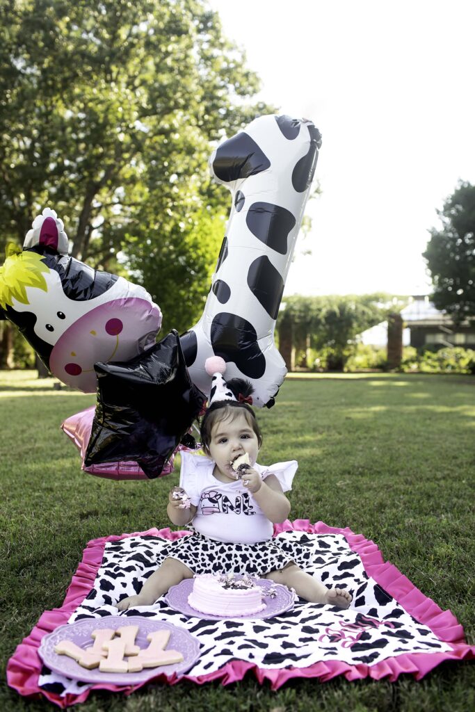 In the peaceful outdoors of Clark Gardens, Mineral Wells, TX, a 1-year-old happily smashes their birthday cake on the grass, creating a fun and memorable moment