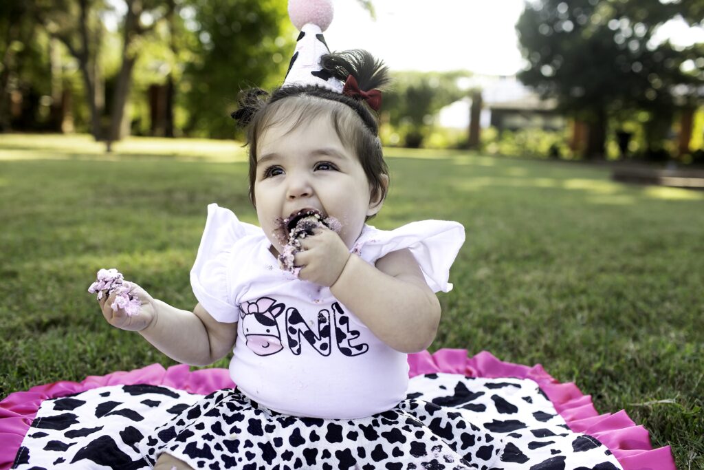 A 1-year-old laughs as they enjoy their cake smash on the grass in Clark Gardens, Mineral Wells, TX, surrounded by nature’s beauty and a stunning garden backdrop