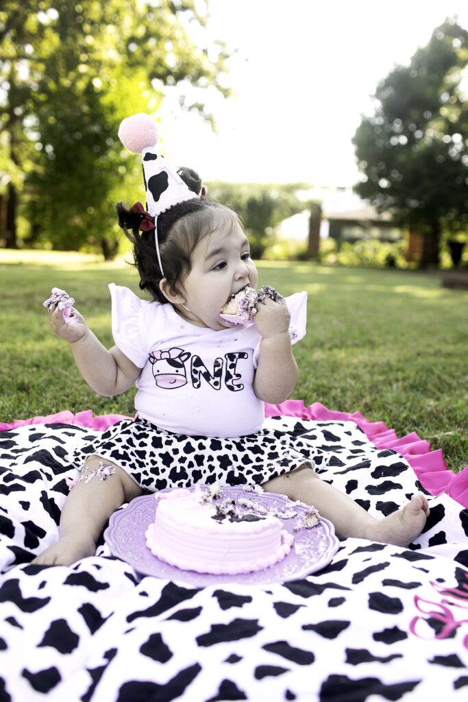 Captured outdoors at Clark Gardens in Mineral Wells, TX, a 1-year-old eagerly digs into their cake during a fun and messy cake smash on the grass