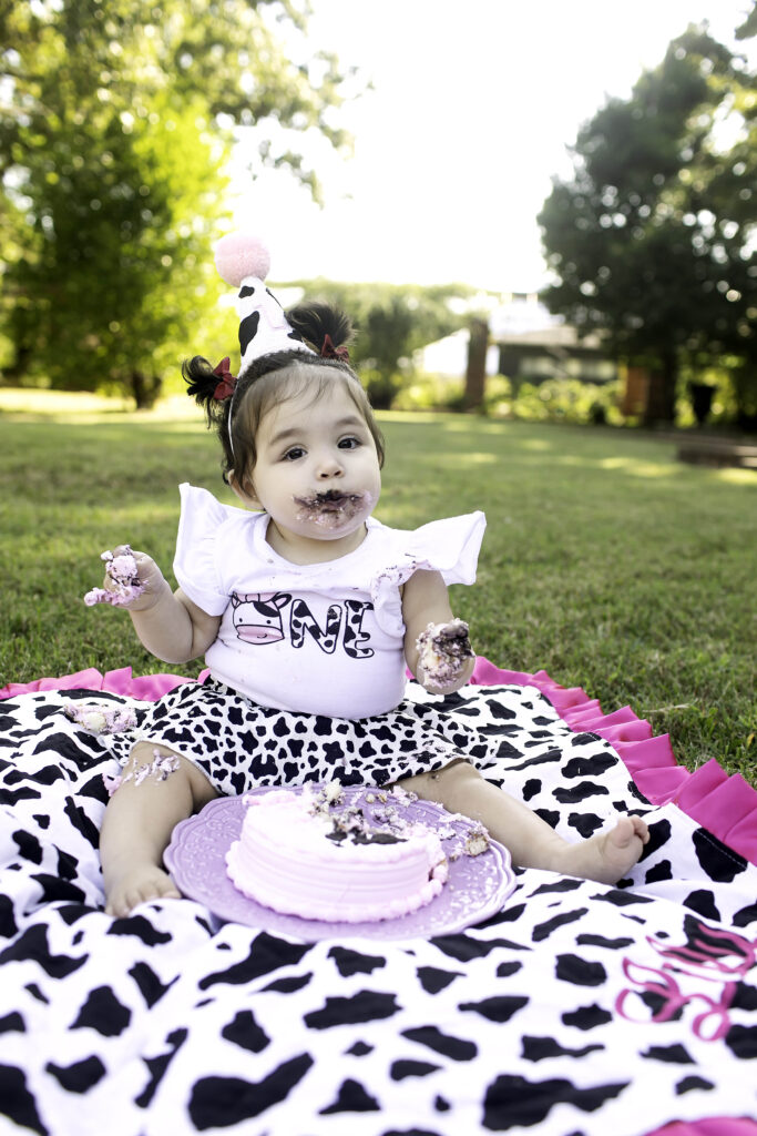 A 1-year-old joyfully smashes their birthday cake on the grass at Clark Gardens in Mineral Wells, TX, surrounded by vibrant greenery and colorful flowers