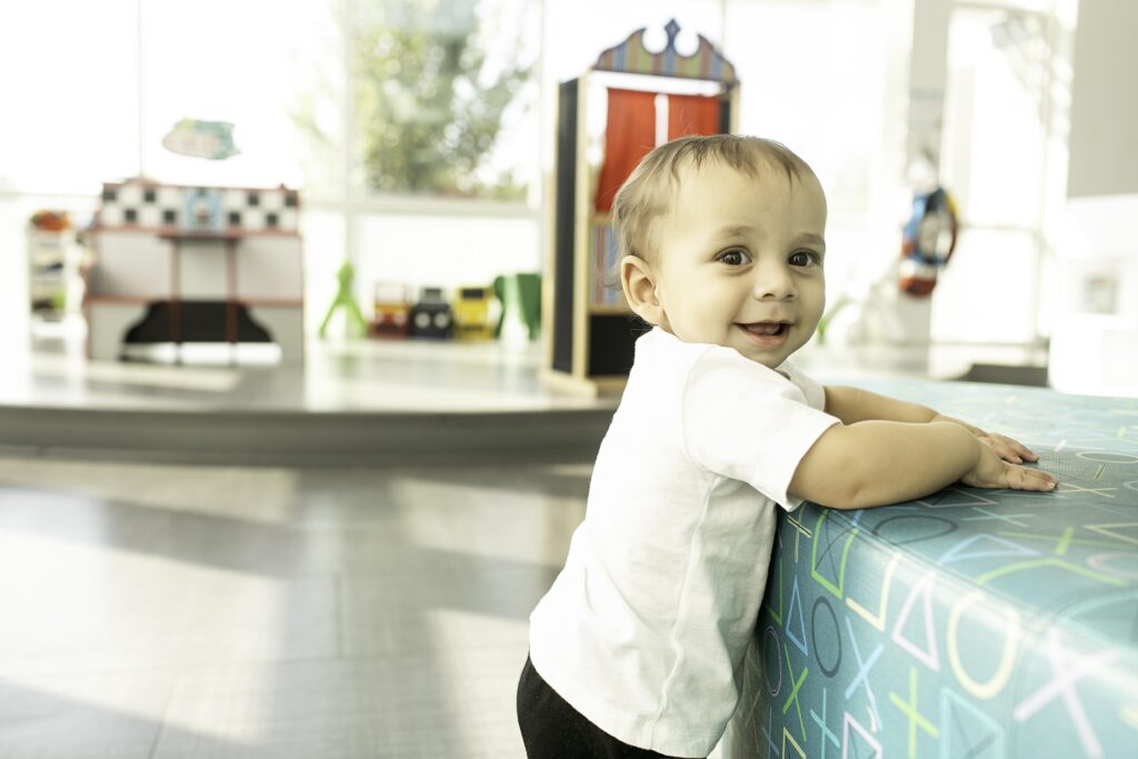 In the Library at Fort Worth, TX, a 1-year-old smiles happily, surrounded by shelves of books, creating a memorable 1st birthday photoshoot