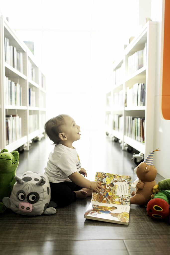 A 1-year-old beams with joy during their birthday photoshoot at the Library in Fort Worth, TX, surrounded by shelves of books in this charming and meaningful setting.