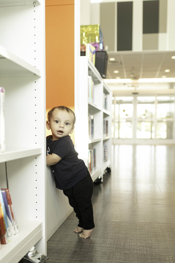 A 1-year-old beams with joy during their birthday photoshoot at the Library in Fort Worth, TX, surrounded by shelves of books in this charming and meaningful setting.