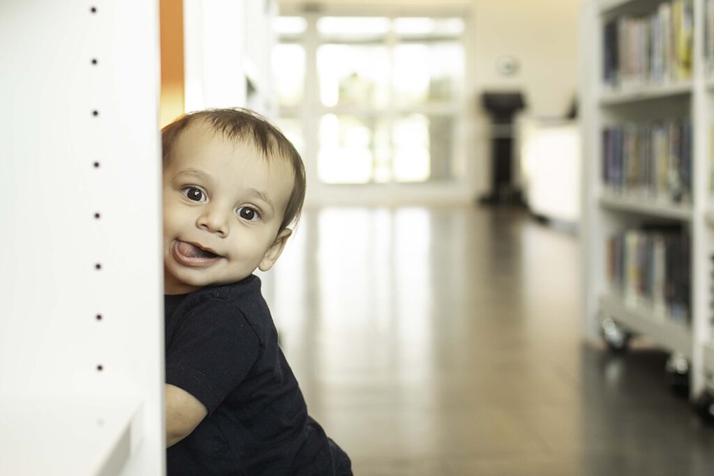 A 1-year-old beams with joy during their birthday photoshoot at the Library in Fort Worth, TX, surrounded by shelves of books in this charming and meaningful setting.