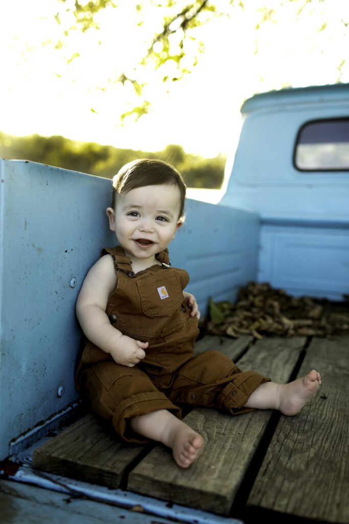 Boy in overalls smiling happily while sitting on the hood of an old blue truck.