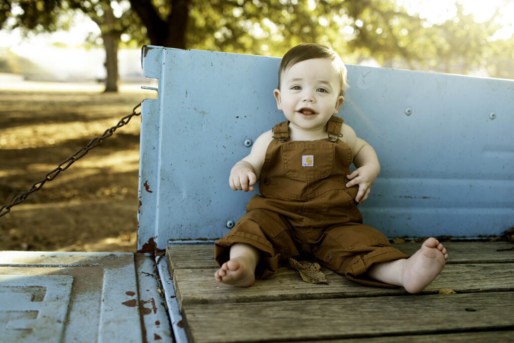 Boy in overalls playfully sitting on the tailgate of an old blue truck, surrounded by rustic charm.