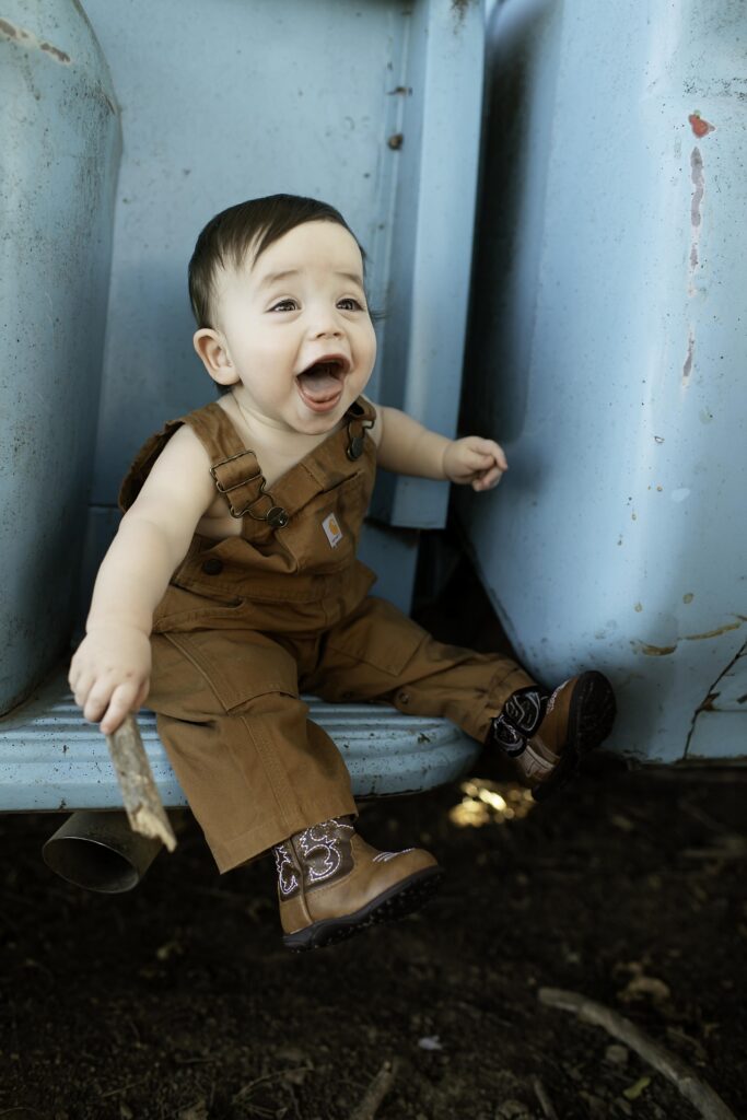 Sweet moment of a boy in overalls sitting on the bumper of an old blue truck, captured in a rustic setting