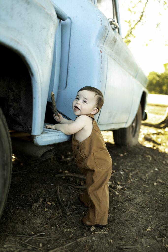 Boy in overalls standing on the step of an old blue truck, gazing into the distance