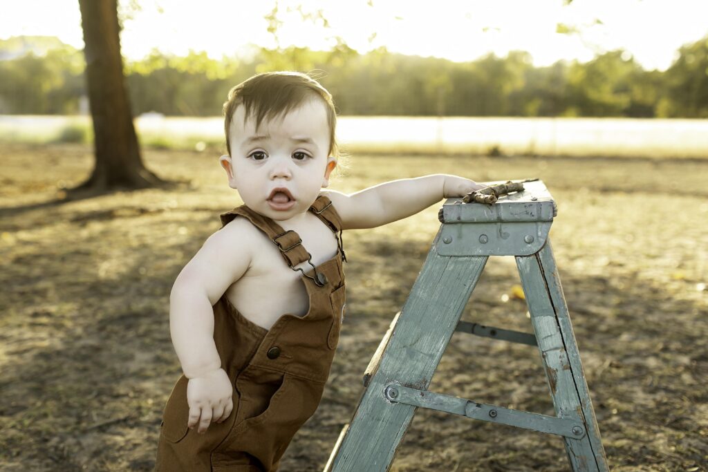 Boy in overalls playfully holding a mini ladder, smiling in a fun outdoor setting