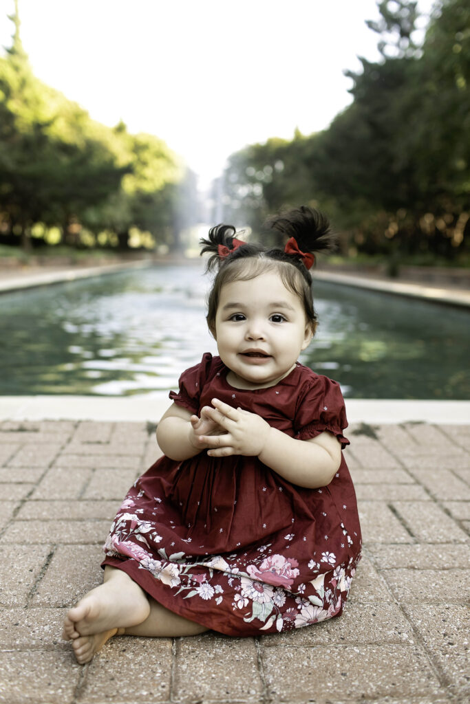 In the peaceful setting of Clark Gardens in Mineral Wells, TX, a 1-year-old giggles joyfully during their first birthday photoshoot, surrounded by vibrant flora.