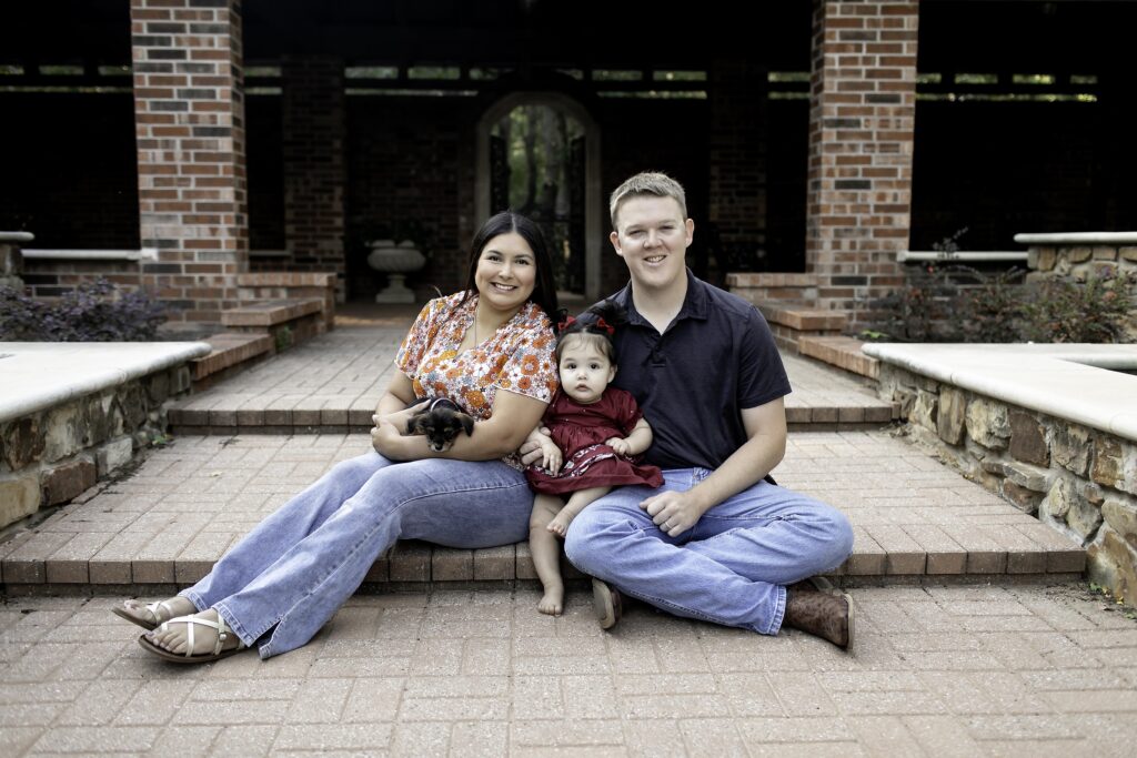  mom and dad smile lovingly at their 1-year-old as they celebrate their first birthday in the beautiful Clark Gardens, Mineral Wells, TX, surrounded by nature