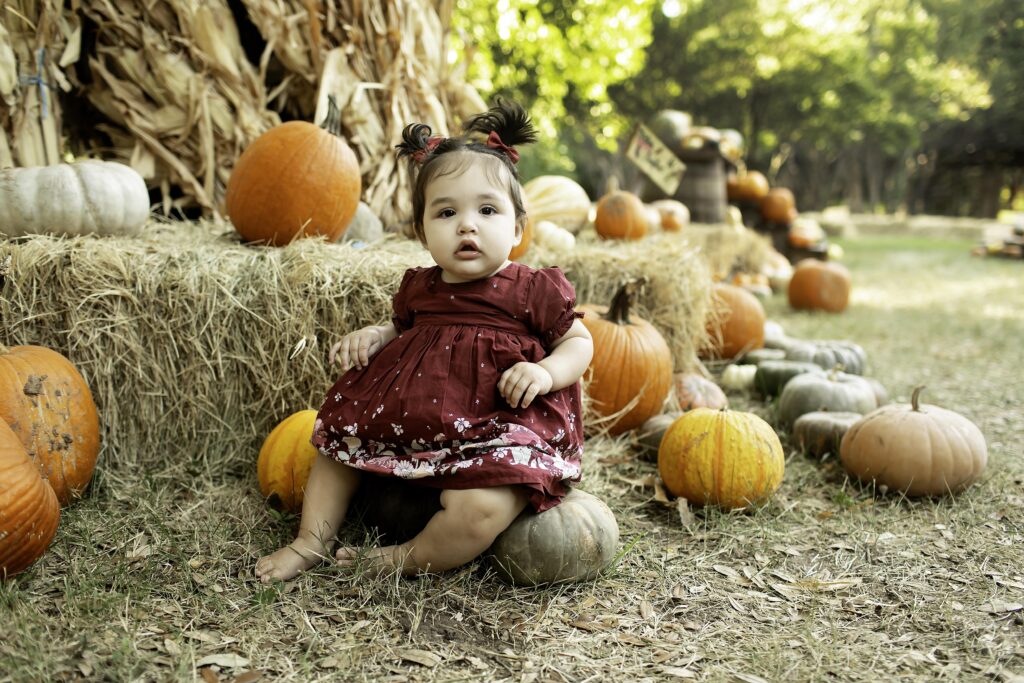 A 1-year-old girl sits among vibrant pumpkins in Clark Gardens, Mineral Wells, TX, celebrating her first birthday with a charming autumn backdrop