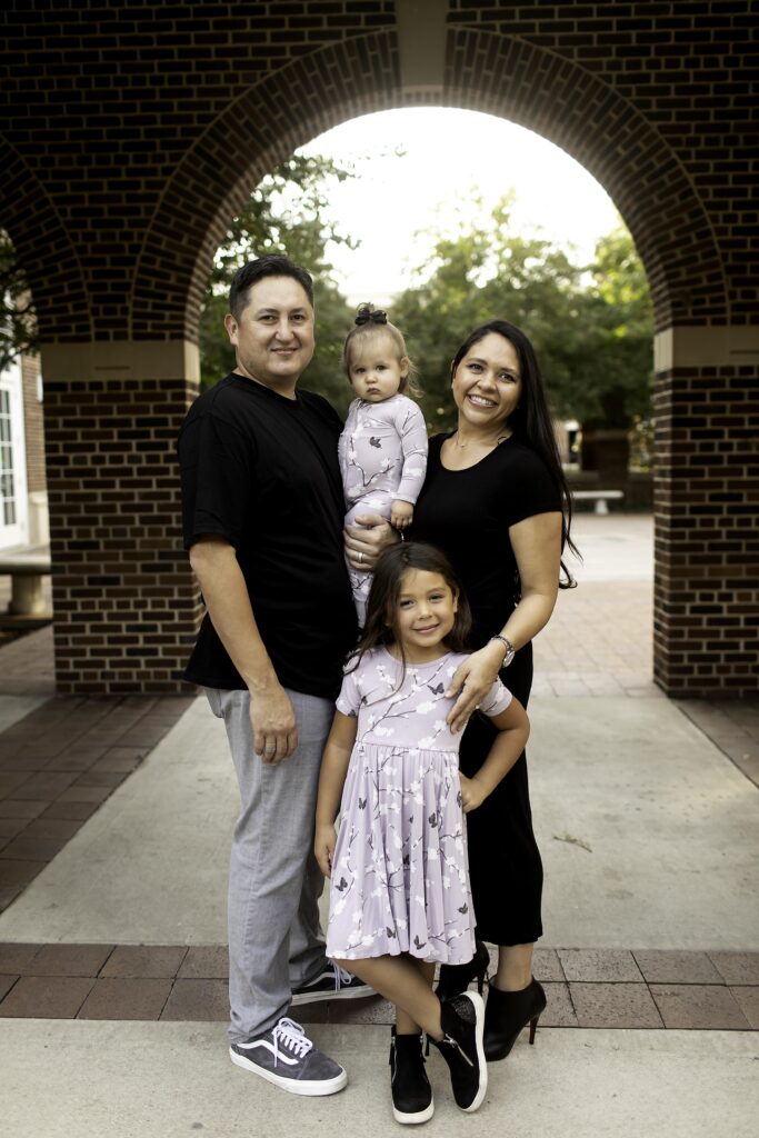 Family portrait session at Southern Methodist University (SMU) in Dallas, Texas, with the family posing against the iconic campus architecture and beautifully manicured landscapes. The group smiles together in front of the historic buildings, with lush greenery and vibrant flowers creating a welcoming, picturesque backdrop. The sunlight bathes the scene, highlighting the family’s connection and the university’s elegant setting