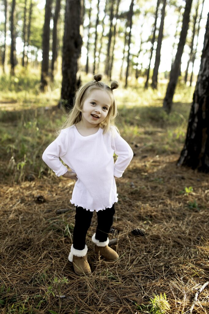 Big sister strikes a sassy pose in the pine forest of Decatur, TX, with a playful smile, exuding confidence and charm amidst nature’s beauty