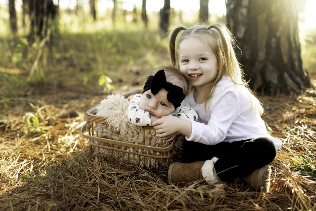 Big sister gently snuggles her 2-month-old sibling, both wrapped in soft blankets and surrounded by the peaceful pine trees of Decatur, TX, sharing a moment of love