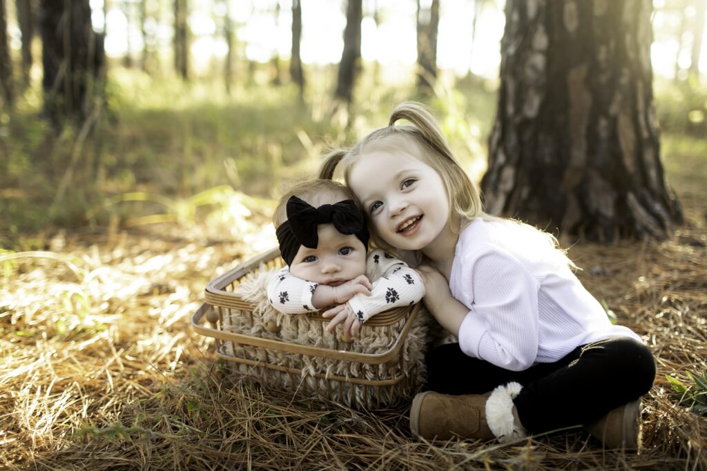 In Decatur’s serene pine forest, big sister lovingly holds her 2-month-old sibling, both snuggled together in a cozy basket, creating a heartwarming family moment.