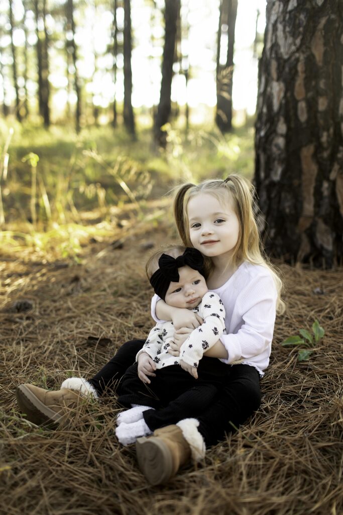 Big sister lovingly snuggles her 2-month-old sibling in the warmth of Decatur's pine trees, sharing a tender moment surrounded by nature's beauty