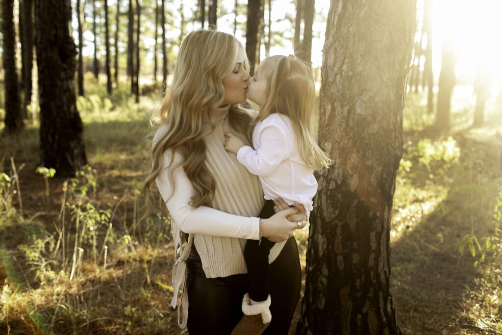 In the peaceful woods of Decatur, a mom kneels down to her 3-year-old, both surrounded by tall pine trees, sharing a moment of love and connection