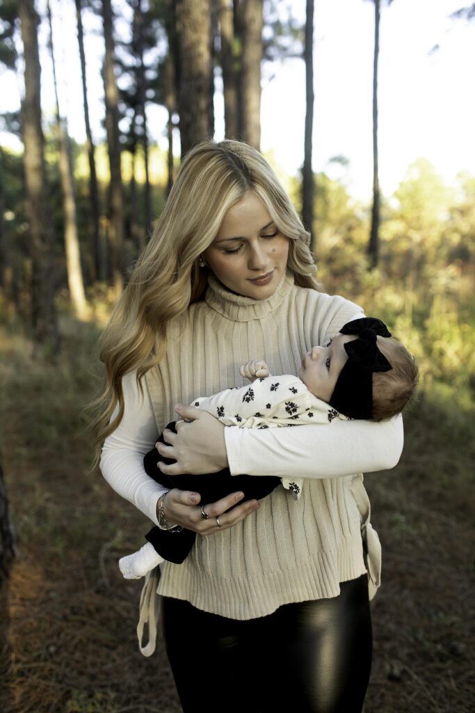 A mother holds her newborn gently in her arms as they stand among the tall, lush pine trees of Decatur, TX, creating a timeless moment of love