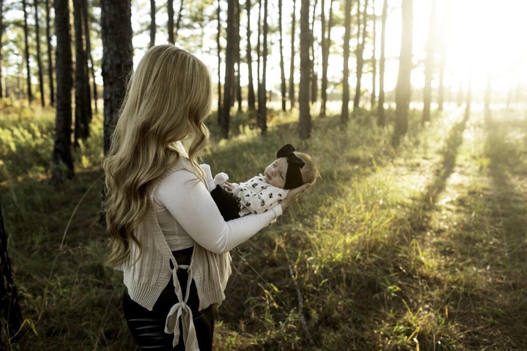 A mom gently holds her newborn in her arms, surrounded by the tall pine trees of Decatur, TX, capturing a peaceful and intimate moment in nature