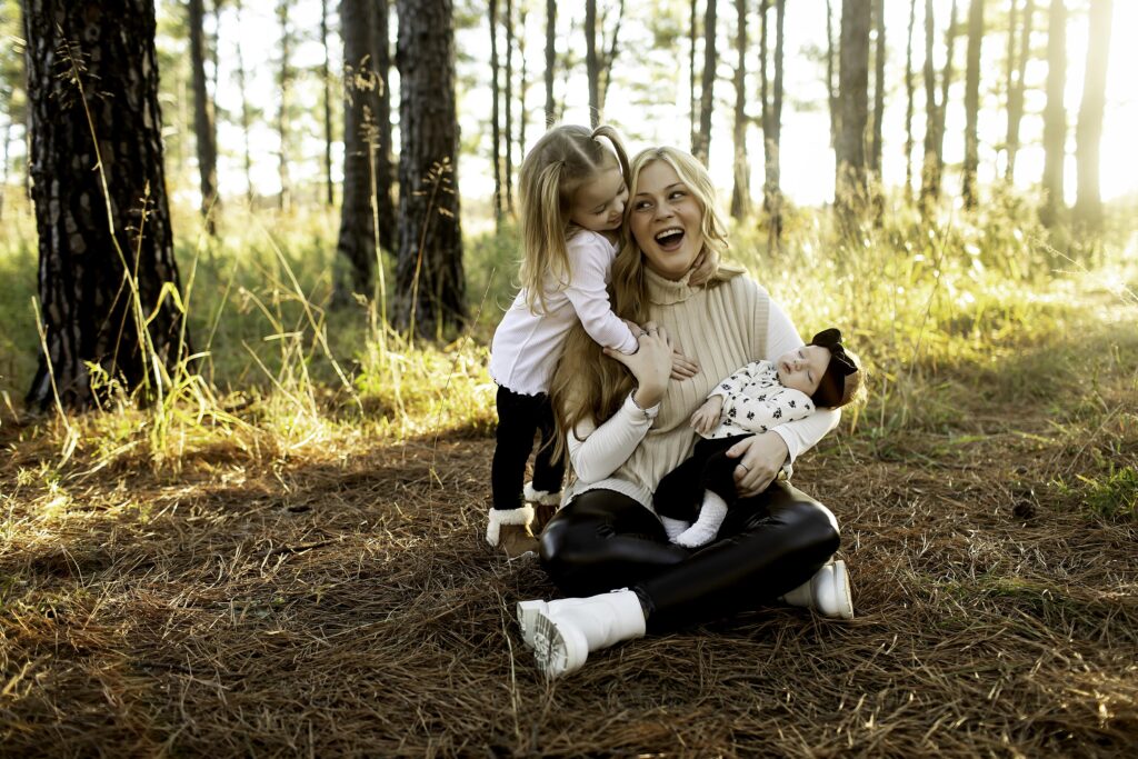 Captured in Decatur’s pine woods, a mom holds her newborn while giving her 3-year-old a loving kiss, both children basking in the warmth of nature’s embrace