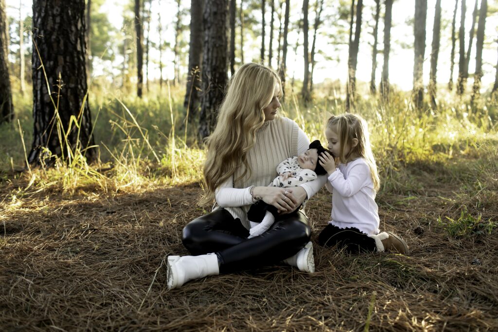 A mother holds her newborn while her 3-year-old happily leans into her for a loving embrace, surrounded by the peaceful pine trees of Decatur, TX