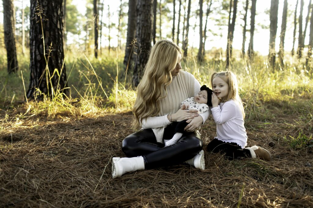 In the serene pine forest of Decatur, a mom tenderly holds her newborn while cuddling with her 3-year-old, creating a heartwarming family scene